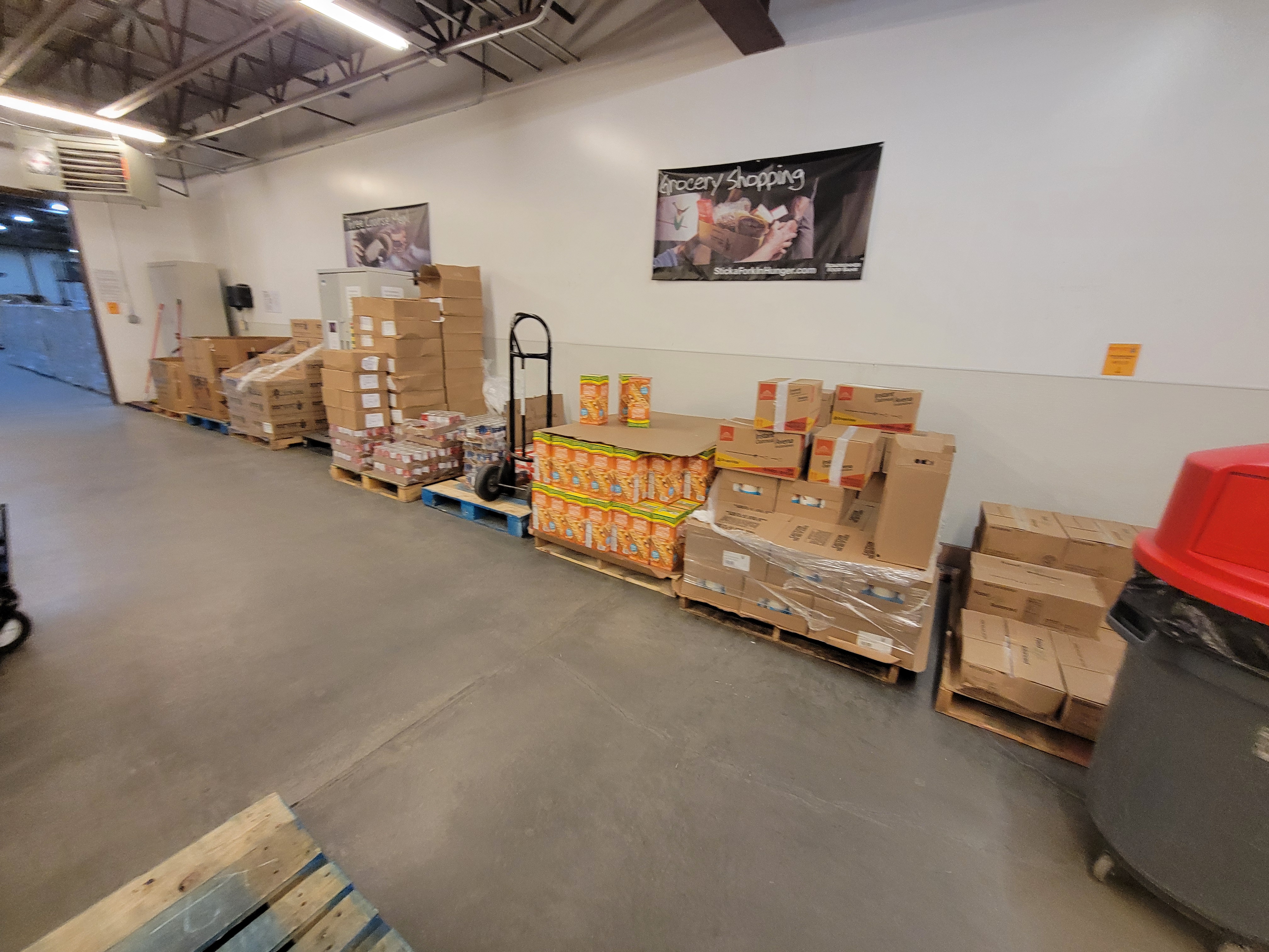 Pallets of food with a trash can near by, and hand truck on top of one.